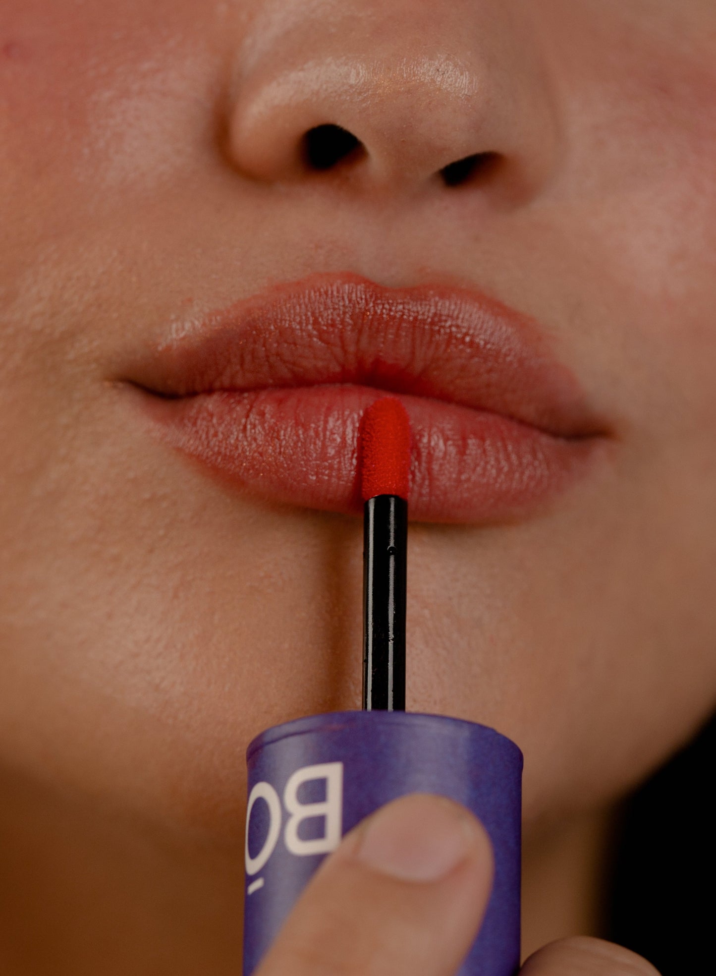 Foto super aproximada de mulher com lábios maquiados com batom líquido lip tint na cor laranja aplicando com aplicador e embalagem roxa com nome BO